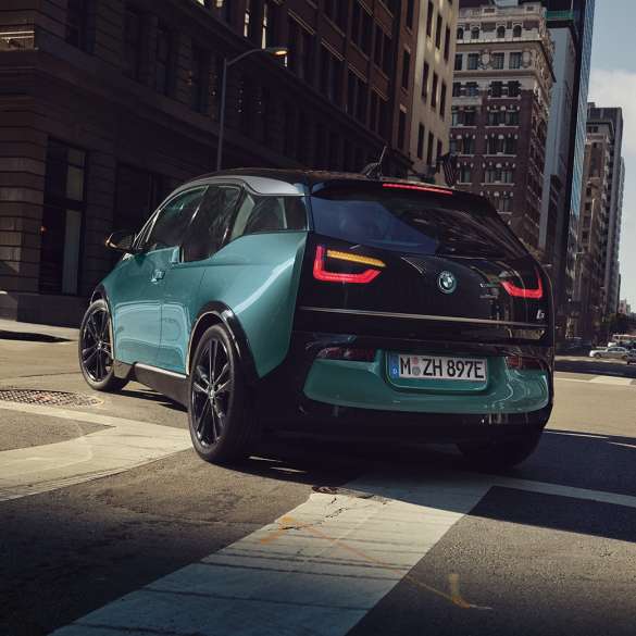 Tročetvrtinski prikaz stražnjeg kraja BMW-a i3 2018 u vožnji Tročetvrtinski prikaz stražnjeg kraja BMW-a i3 I01 2018 Jucaro bež boje s Frozen Grey metalik detaljima u vožnji u urbanom okruženju