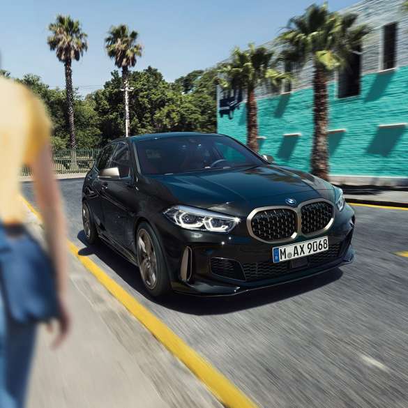 BMW M135i xDrive, tročetvrtinski prikaz prednjeg kraja. BMW M135i xDrive u Black Sapphire metalik boji, tročetvrtinski prikaz prednjeg kraja prilikom vožnje, sa ženom u prvom planu.