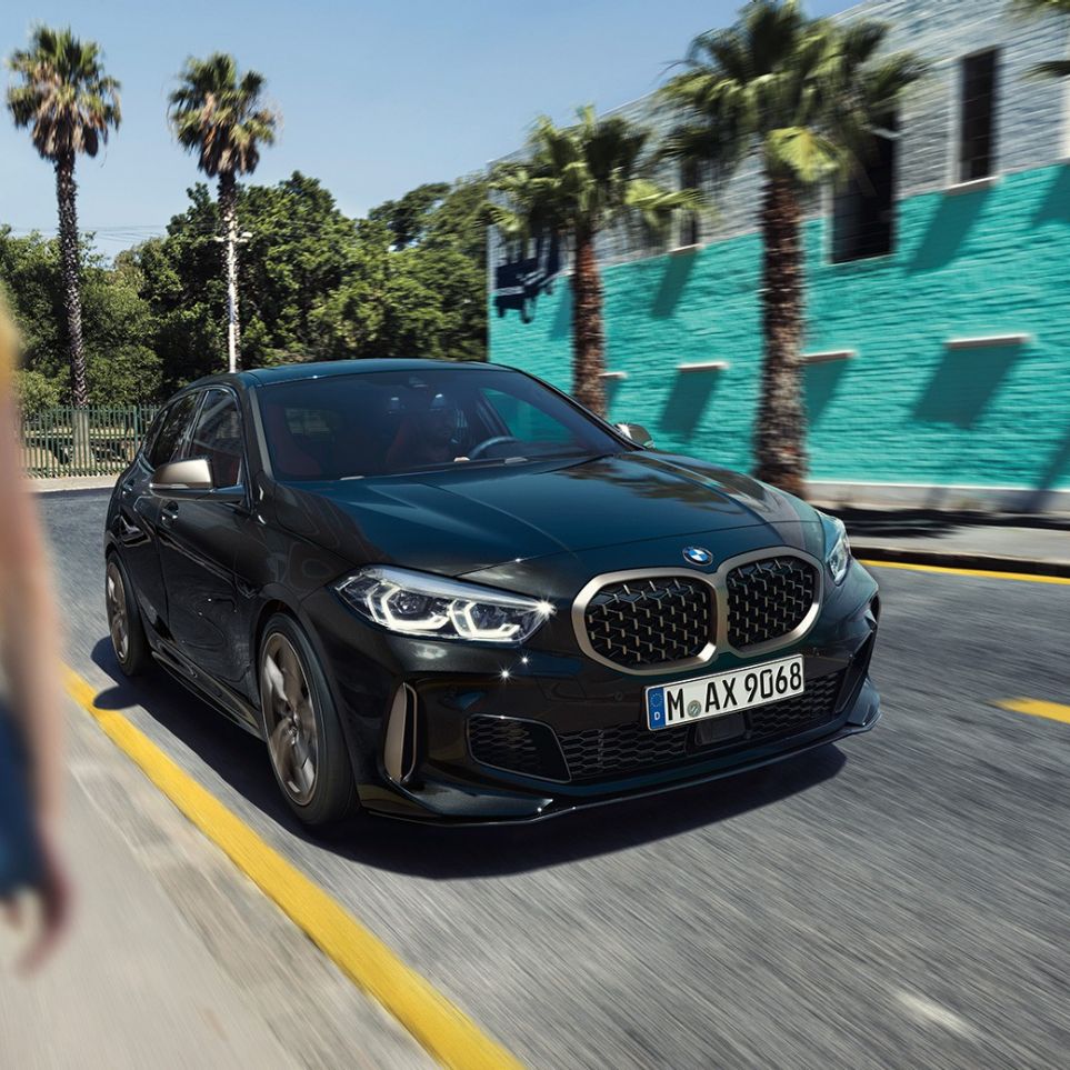 BMW M135i xDrive, tročetvrtinski prikaz prednjeg kraja. BMW M135i xDrive u Black Sapphire metalik boji, tročetvrtinski prikaz prednjeg kraja prilikom vožnje, sa ženom u prvom planu.