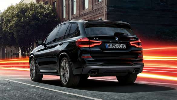 BMW X3 M40i, tročetvrtinski stražnji prikaz. BMW X3 M40i u Black Sapphire metalik boji, u vožnji, vanjština, tročetvrtinski stražnji prikaz ispred urbane pozadine sa svijetlosnim trakama.