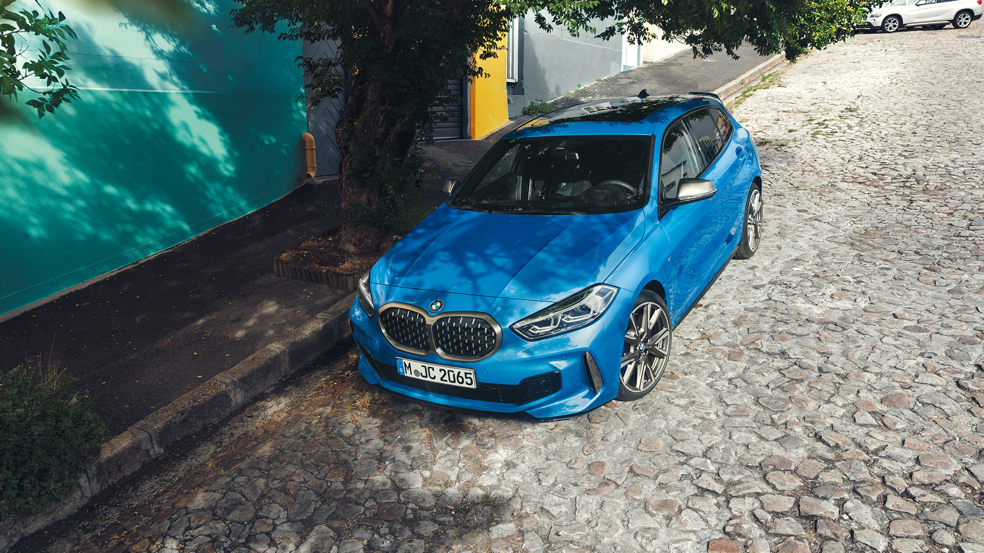 BMW serije 1 sa strane ceste. BMW serije 1 u Misano Blue metalik boji, tročevrtinski prikaz prednjeg kraja, prikaz modela parkiranog sa strane ceste iz ptičje perspektive.