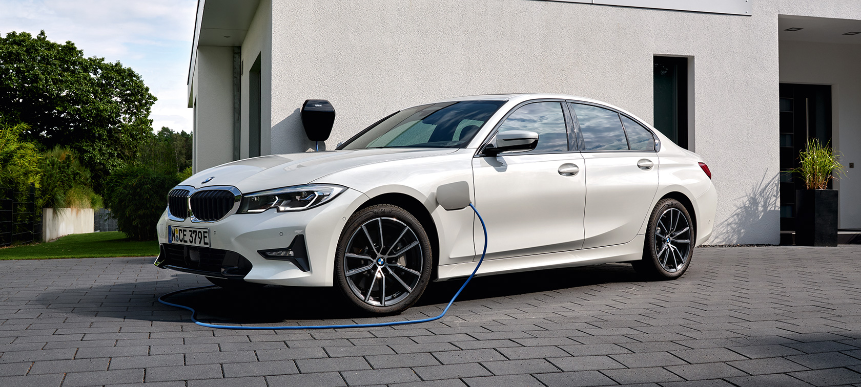 Tročetvrtinski bočni prikaz plug-in hibrida BMW-a serije 3 Limuzina 2021 G20 na napajanju Tročetvrtinski bočni prikaz plug-in hibrida BMW-a serije 3 Limuzina 2021 G20 na napajanju ispred garaže