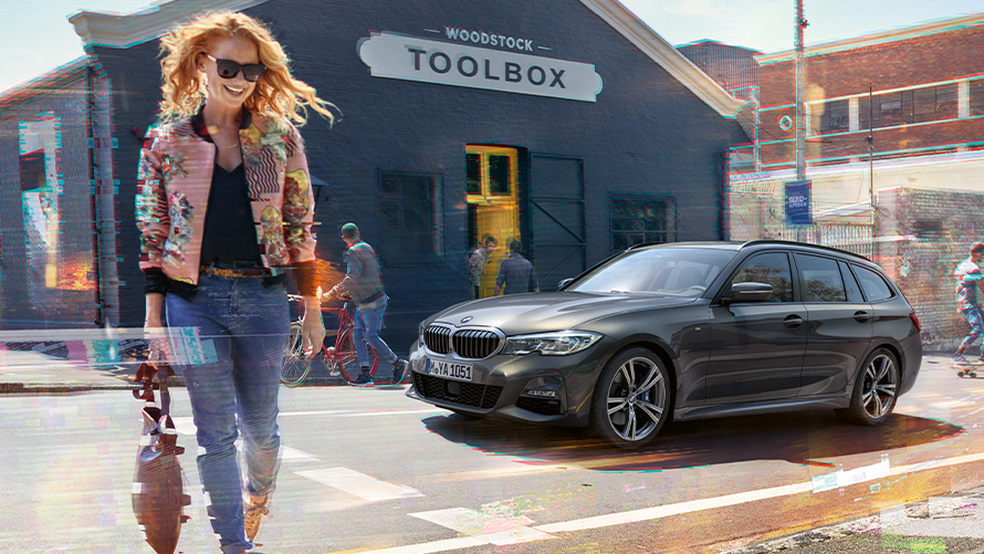 BMW serije 3 Touring, (G21) tročetvrtinski pogled na ženu ispred drvne radionice. Žena prelazi ulicu ispred BMW-a serije 3 Touring (G21) ispred drvne radionice.