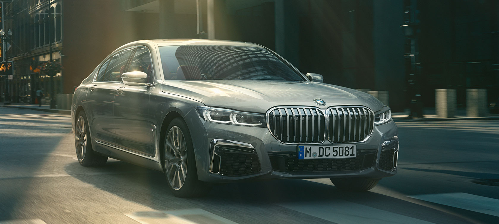 Sivi BMW serije 7 Limuzina u vožnji, tročetvrtinski prikaz Sivi BMW serije 7 Limuzina u vožnji kroz urbano okruženje u tročetvrtinskom prednjem prikazu