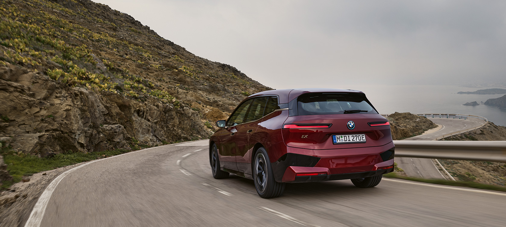 BMW iX i20 2021 električni SUV; Prikaz BMW-a iX xDrive50 u vožnji priobalnom cestom BMW iX i20 2021 električni SUV; Prikaz stražnjeg kraja BMW-a iX xDrive50 Aventurin Red boje u vožnji priobalnom cestom