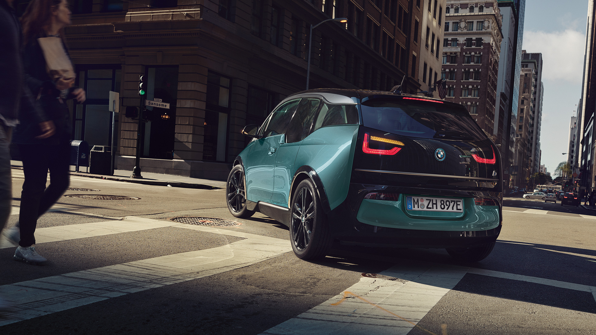Tročetvrtinski prikaz stražnjeg kraja BMW-a i3 2018 u vožnji Tročetvrtinski prikaz stražnjeg kraja BMW-a i3 I01 2018 Jucaro bež boje s Frozen Grey metalik detaljima u vožnji u urbanom okruženju