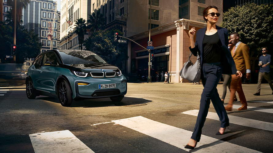 BMW i3 I01 tročetvrtinski pogled sprijeda sa ženom koja prelazi cestu preko pješačkog prijelaza BMW i3 I01 Blue Ridge Mountain metalik sa Frozen Grey metalik naglascima tročetvrtinski pogled sprijeda sa ženom koja prelazi cestu preko pješačkog prijelaza