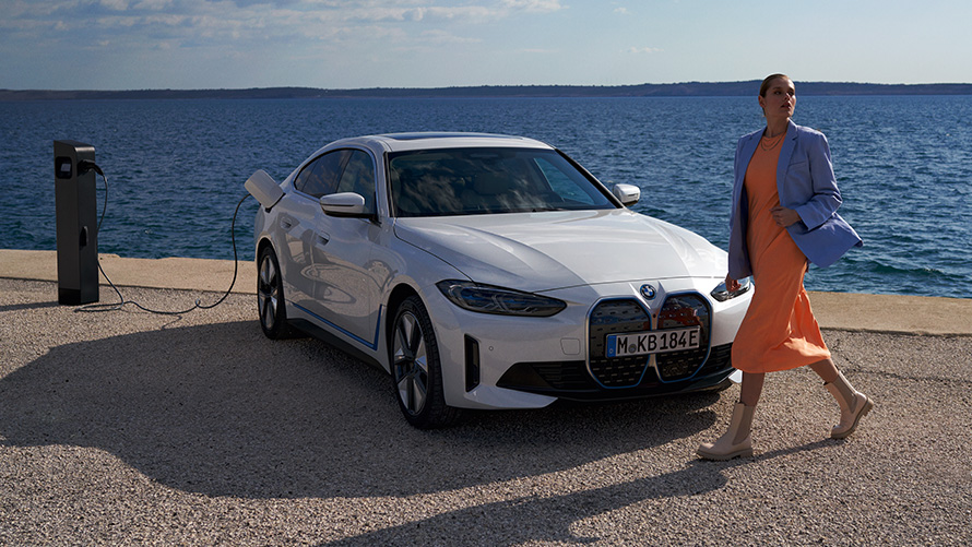 Tročetvrtinski pogled sprijeda BMW i4 eDrive40 G26 2021 na stanici za punjenje Žena ispred BMW i4 eDrive40 G26 2021 Mineral White tročetvrtinski pogled sprijeda na stanici za punjenje ispred jezera