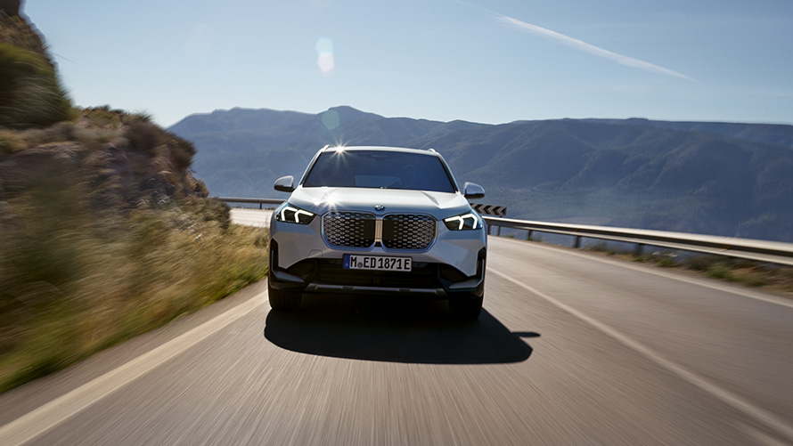 BMW iX1 U11 BEV 2022 pogled sprijeda BMW iX1 U11 BEV 2022 Mineral White metalik pogled sprijeda planinska cesta u vožnji