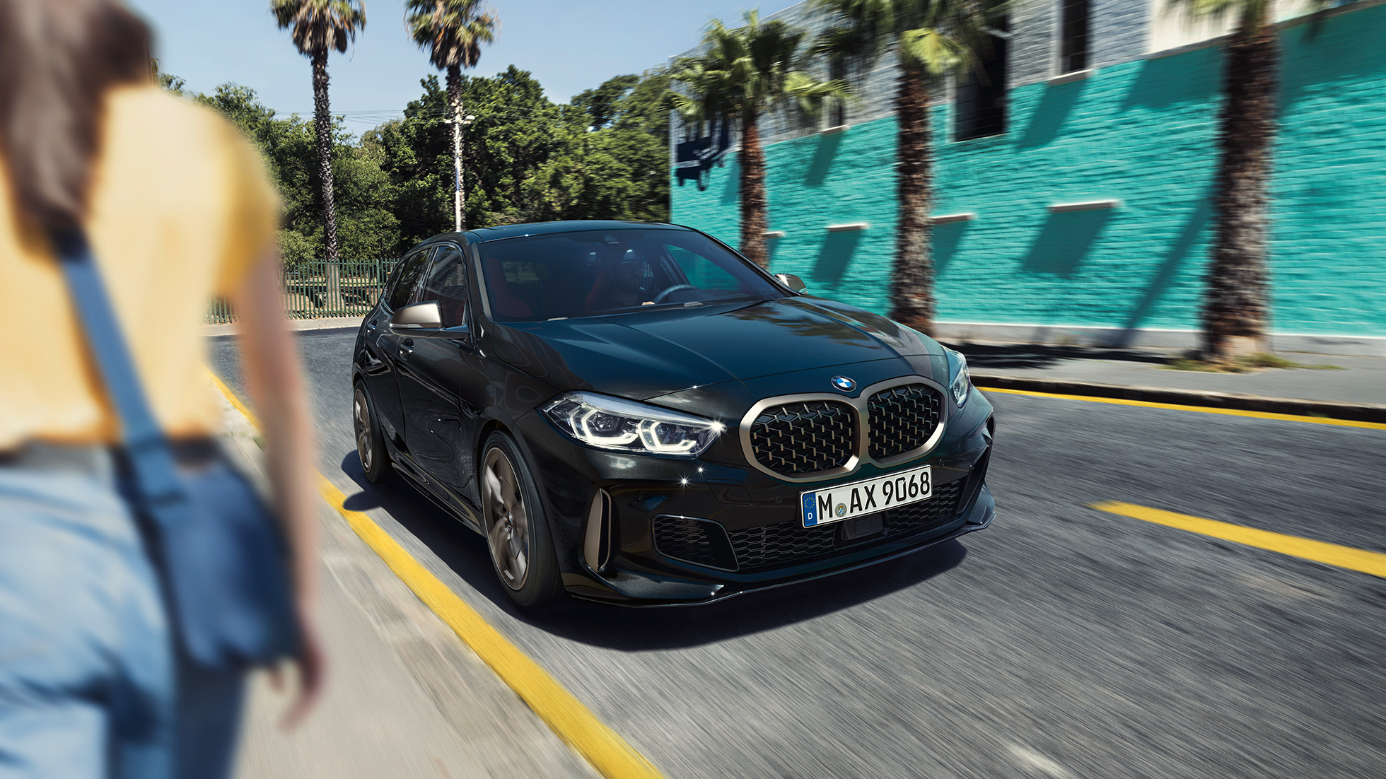 BMW M135i xDrive, tročetvrtinski prikaz prednjeg kraja. BMW M135i xDrive u Black Sapphire metalik boji, tročetvrtinski prikaz prednjeg kraja prilikom vožnje, sa ženom u prvom planu.