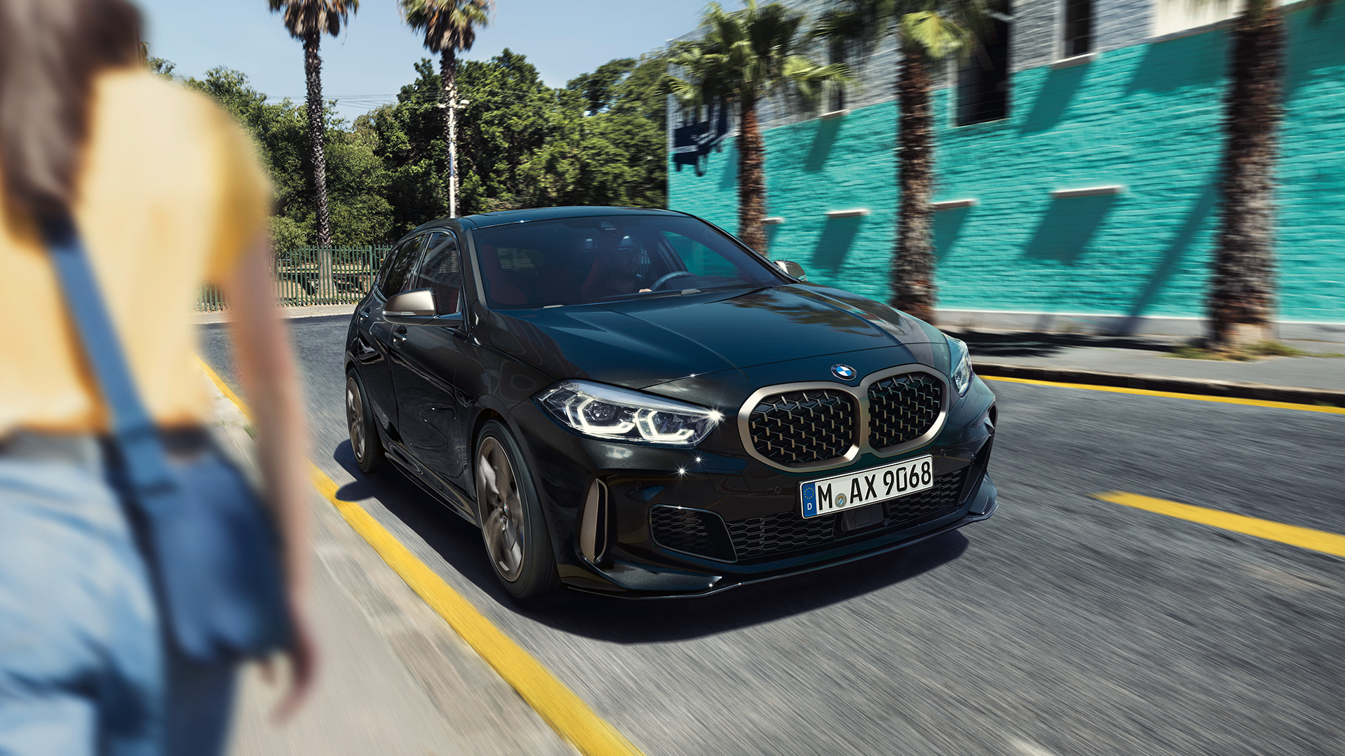 BMW M135i xDrive, tročetvrtinski prikaz prednjeg kraja. BMW M135i xDrive u Black Sapphire metalik boji, tročetvrtinski prikaz prednjeg kraja prilikom vožnje, sa ženom u prvom planu.