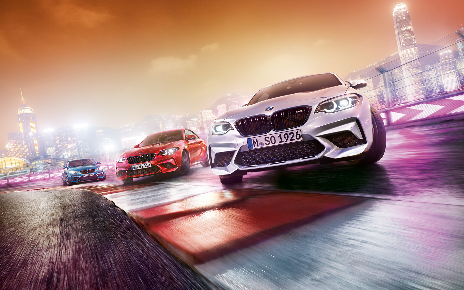BMW M2 Competition u akciji Tri modela BMW M2 Competition na trkaćoj stazi ispred noćnog pejzaža grada.