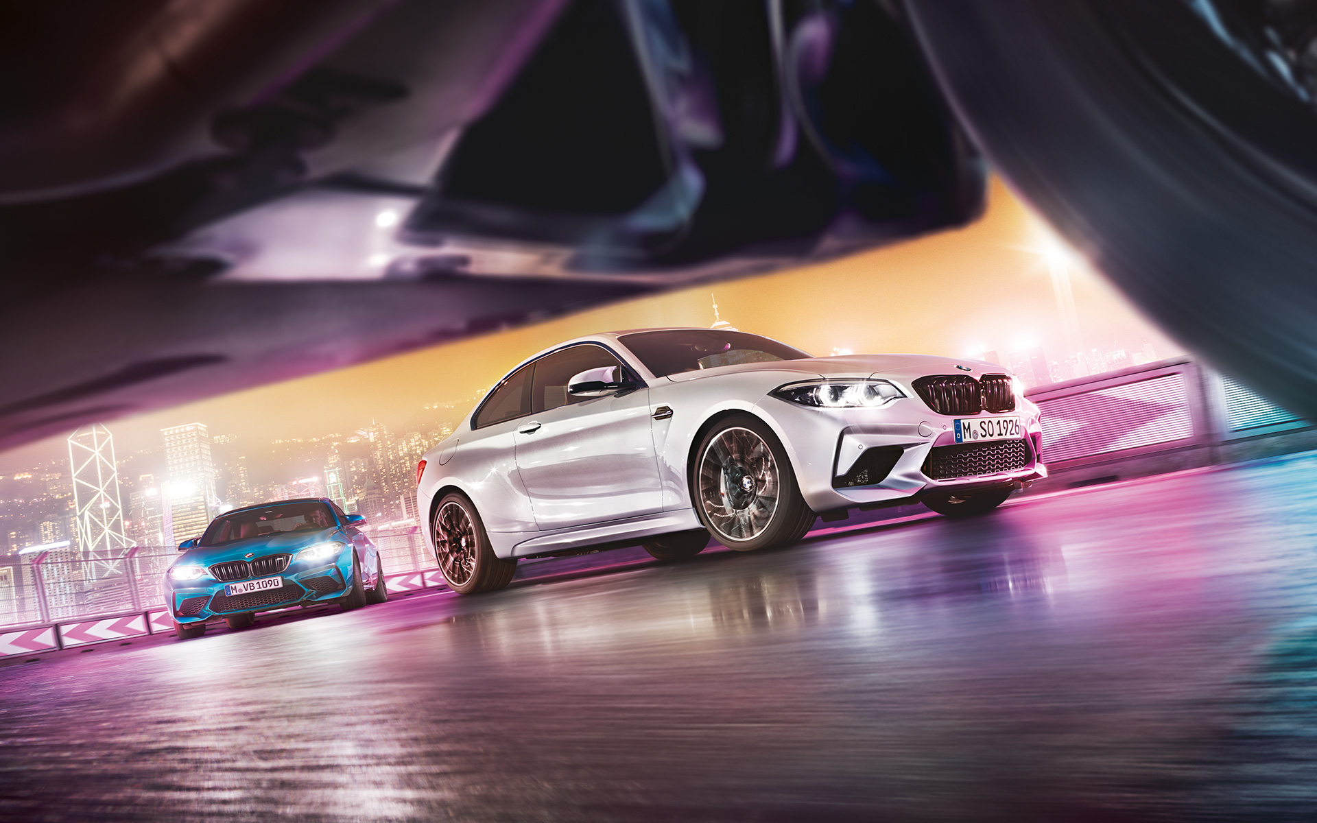 BMW M2 Competition na asfaltu. Dva modela BMW M2 Competition na trkaćoj stazi ispred noćnog pejzaža grada.