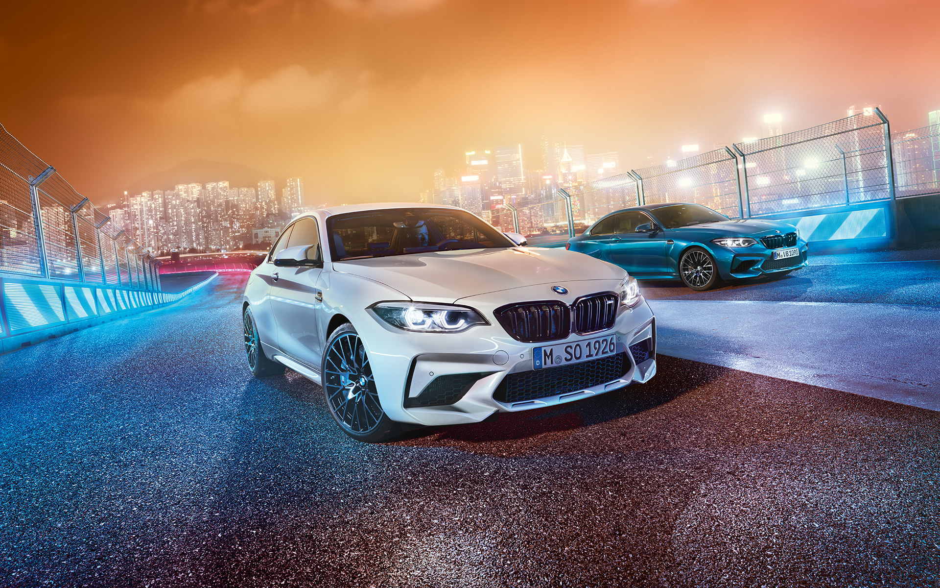 Dva modela BMW M2 Competition na trkaćoj stazi Bijeli i plavi modeli BMW M2 Competition ispred noćnog pejzaža grada.