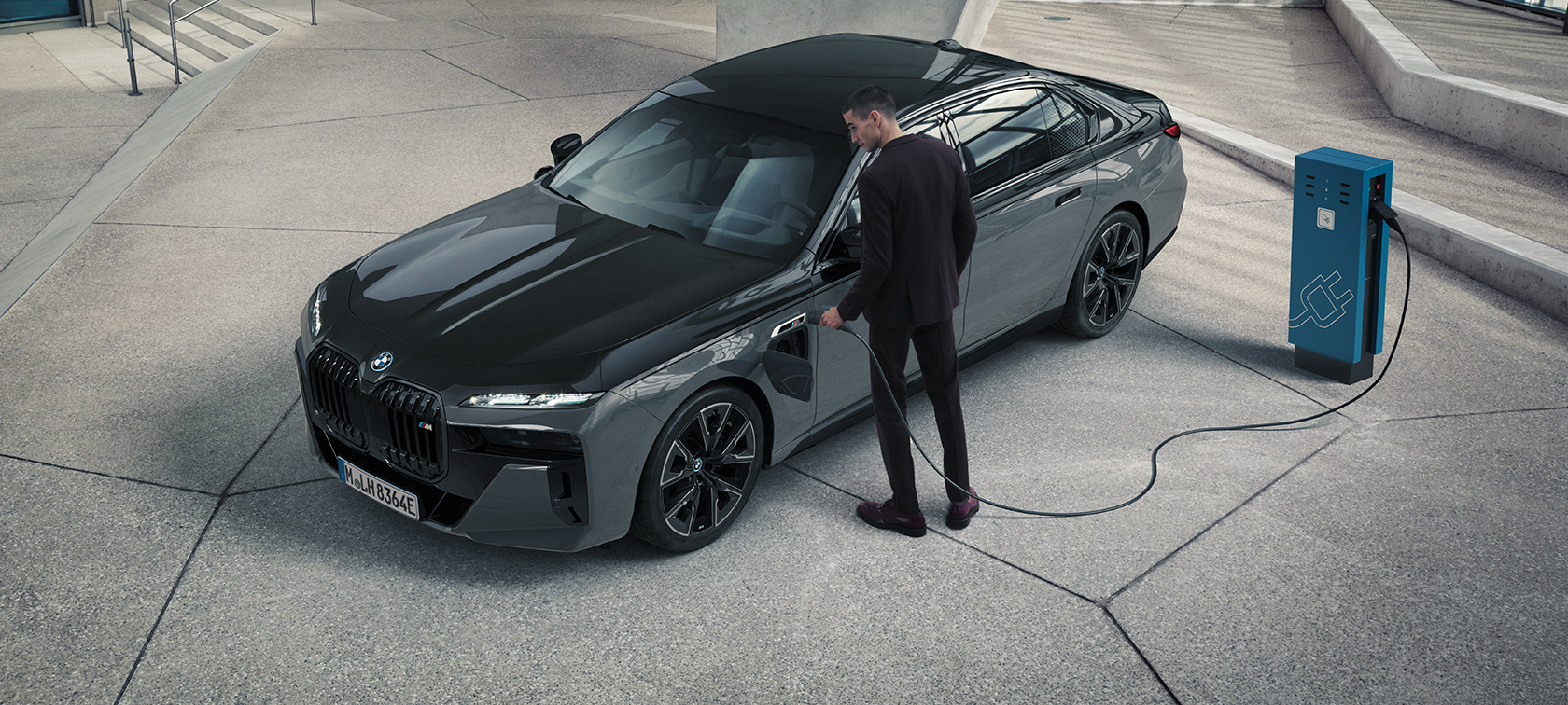 BMW M760e xDrive Limuzina G70 M Performance paket BMW M760e xDrive Limuzina G70 M Performance paket BMW Individual dvotonska Black Sapphire metalik boja BMW Individual Dravite Grey metalik tročetvrtinski prednji prikaz muškarac s kablom za punjenje pokraj stanice za punjenje