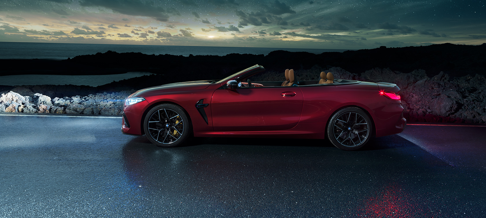 BMW M8 Competition Cabrio, bočni prikaz. BMW M8 Competition Cabrio u Motegi Red metalik boji sa spuštenim mekim krovom, bočni prikaz, na obalnoj cesti pod zvjezdanim nebom.