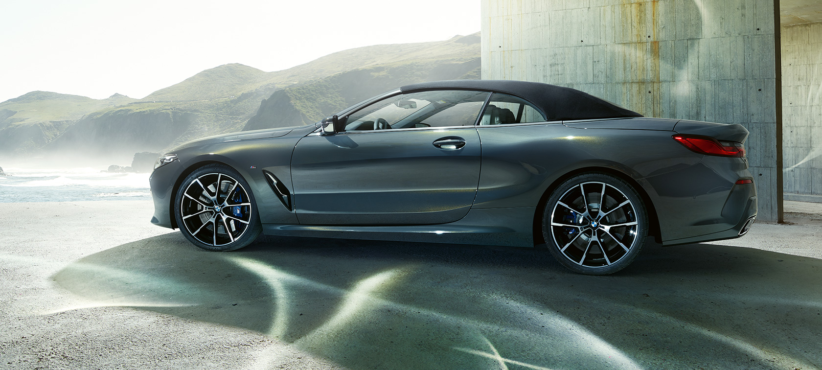 BMW M850i xDrive Cabrio, bočni prikaz. BMW M850i xDrive Cabrio u Dravit Grey metalik boji, s dignutim mekim krovom, bočni prikaz ispred brda.