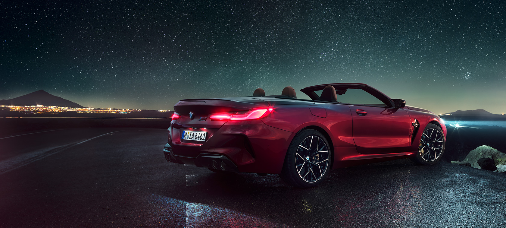 BMW M8 Competition Cabrio, tročetvrtinski prikaz stražnjeg kraja. BMW M8 Competition Cabrio u Motegi Red metalik boji sa spuštenim krovom, tročetvrtinski prikaz stražnjeg kraja, ispod zvjezdanog neba.