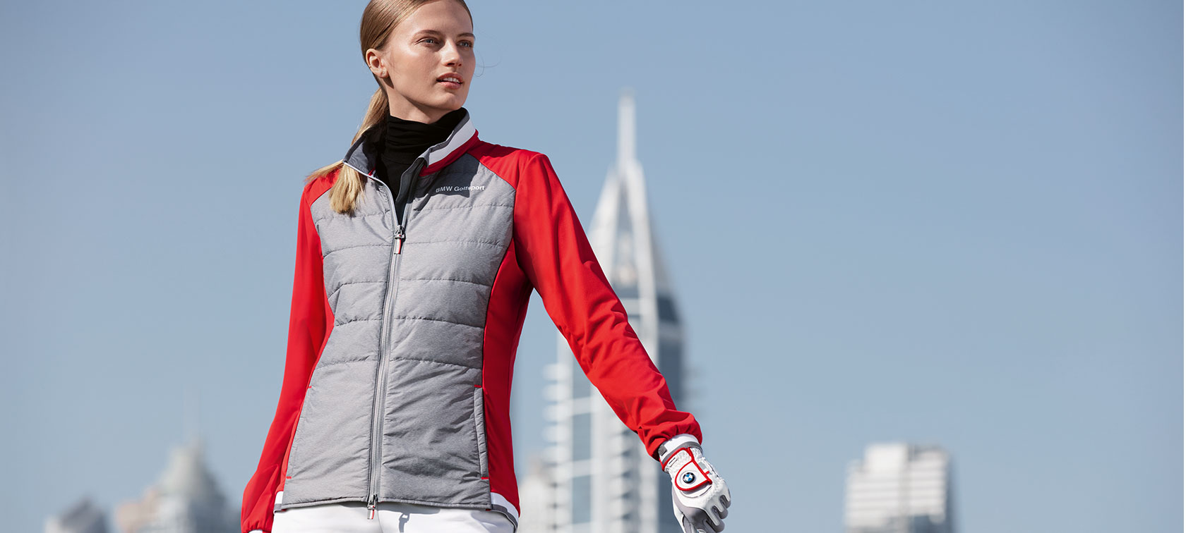 BMW Golfsport ženska jakna. Mlada žena na slici predstavlja vrhunske predmete iz BMW Golfsport kolekcije uz pozadinski obris velikog grada.