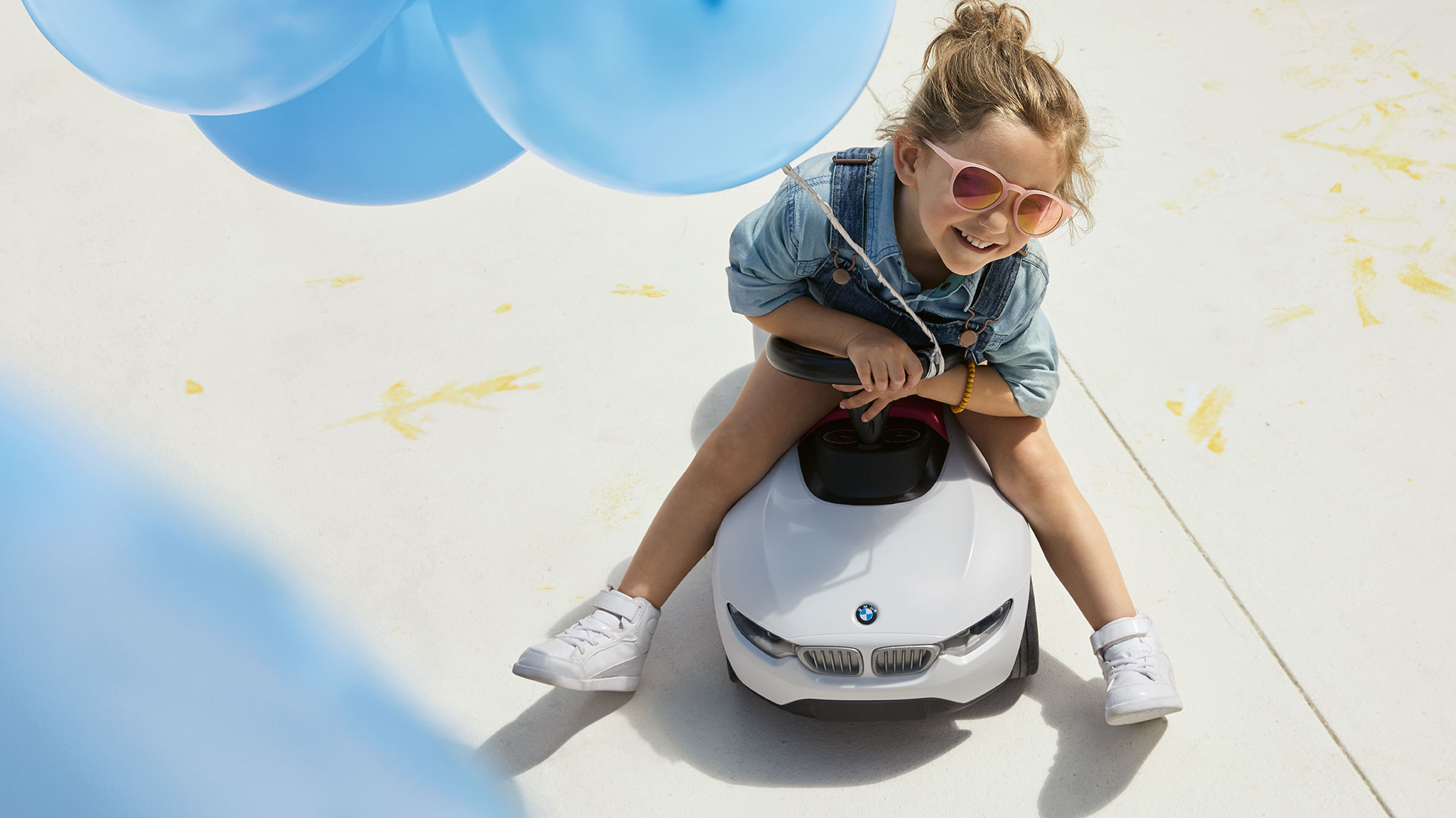 BMW Kids kolekcija. Djevojka s ružičastim sunčanim naočalama i plavim balonom sretno sjedi na BMW Baby Racer igrački.