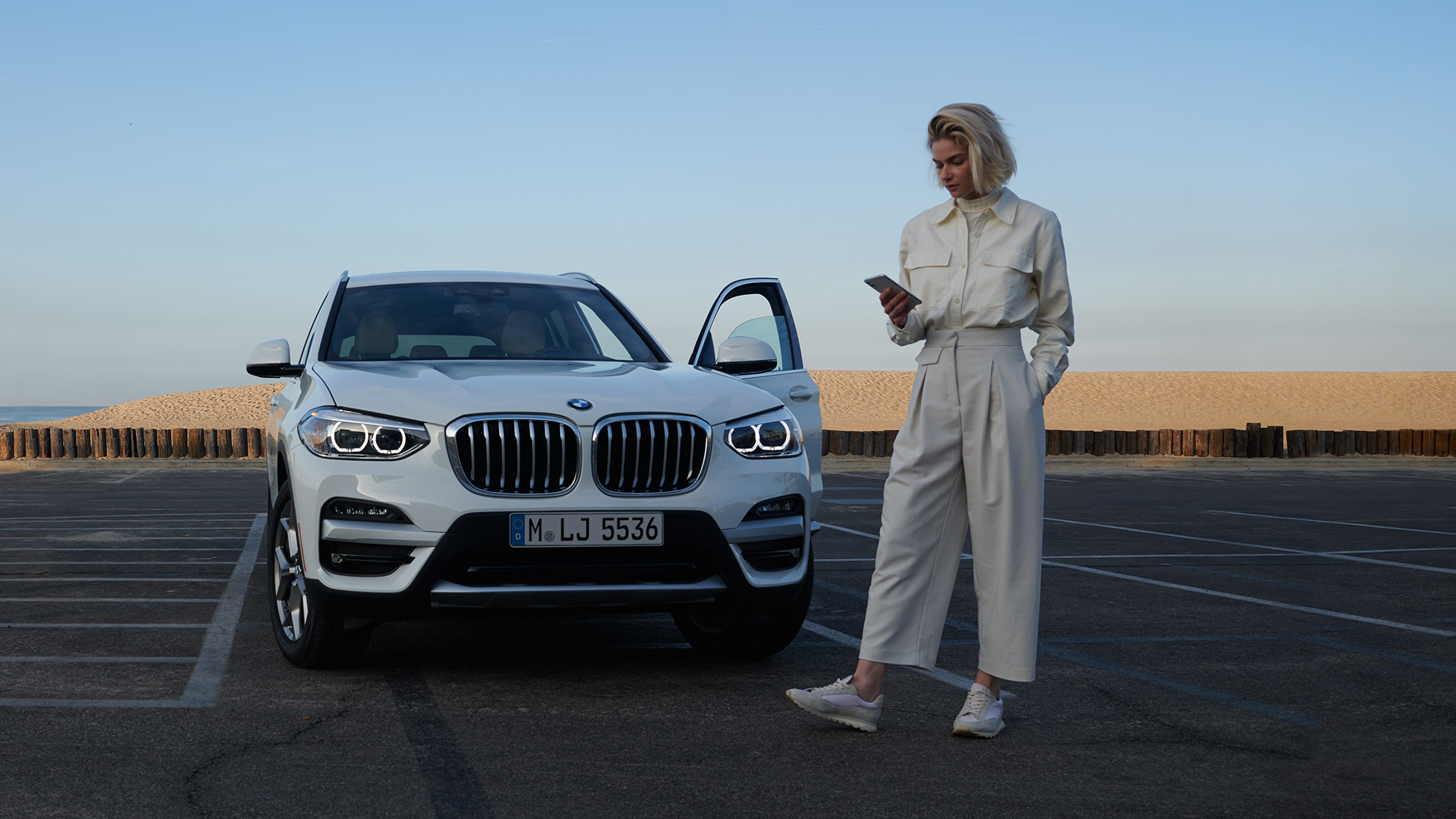 Ženska osoba koristi My BMW aplikaciju na pametnom telefonu Ženska osoba koristi My BMW aplikaciju iz BMW ConnectedDrive-a na pametnom telefonu ispred bijelog BMW X5