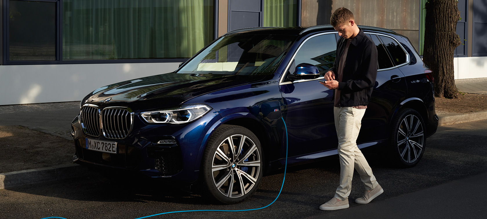 Muškarac koristi My BMW aplikaciju na pametnom telefonu Muškarac koristi My BMW aplikaciju iz BMW ConnectedDrive na pametnom telefonu dok puni svoj BMW X5 Plug-in hibrid