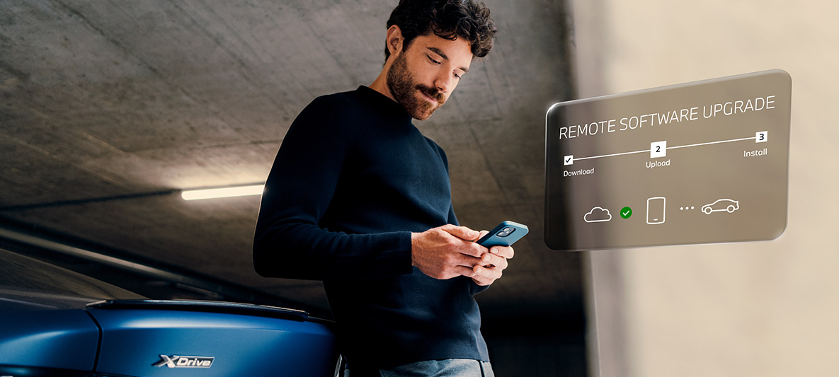 Muškarac se naslanja na BMW straga i gleda u svoj pametni telefon Remote Software Upgrade Muškarac se naslanja na BMW straga i gleda u svoj pametni telefon